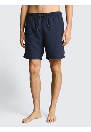 Männlicher Oberkörper und Beine, die dunkelblaue Badehose mit Kordelzug tragen, barfuß auf hellem grauem Hintergrund stehend.