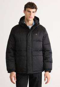 Casaco puffer preto com capuz, apresentando textura acolchoada, fecho de botões, dois bolsos frontais e um logótipo no peito.