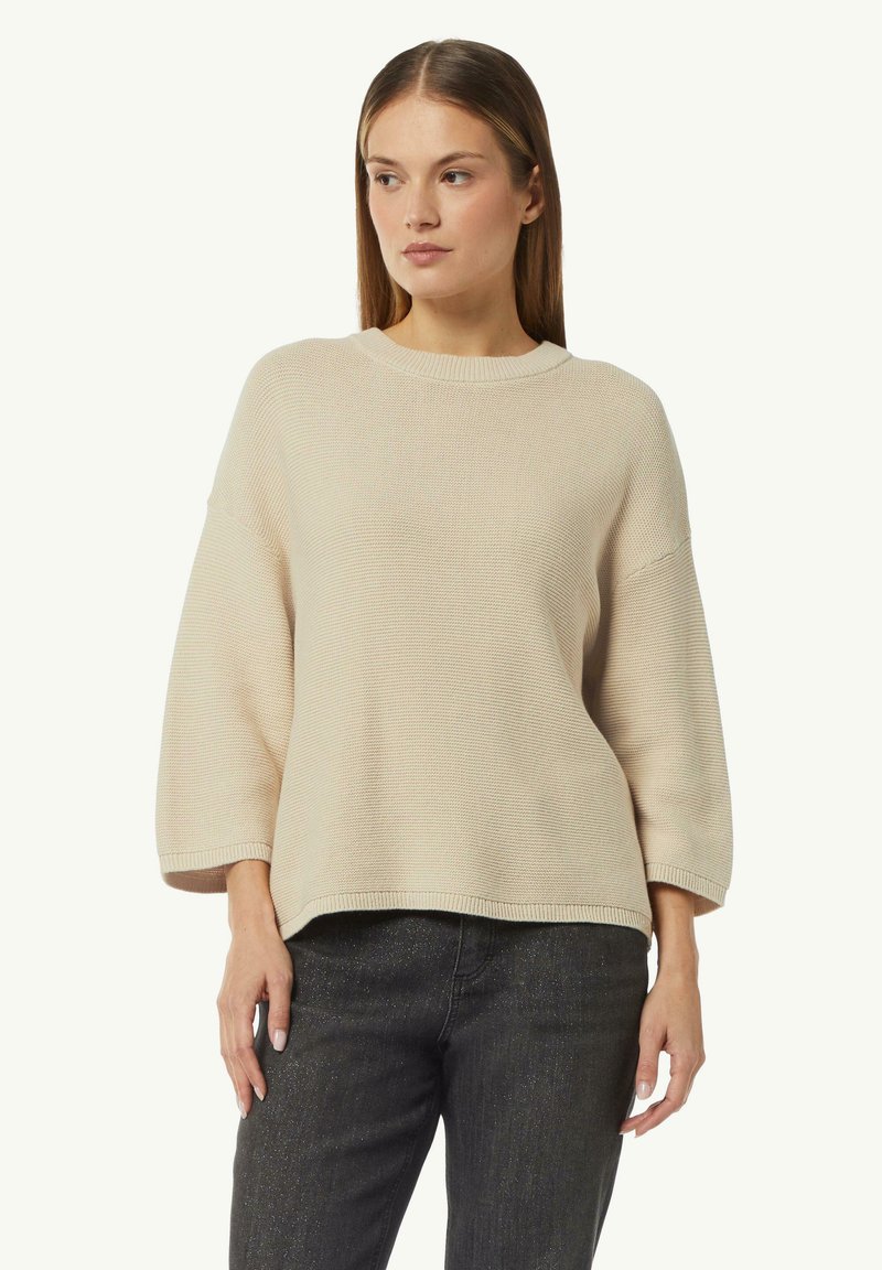Beige gestrickter Pullover mit rundem Halsausschnitt und weiten, kurzen Ärmeln. Strukturierter Stoff mit lässiger Passform, kombiniert mit dunklen Jeans.