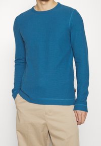 Blå långärmad tröja med rund hals i ribbad struktur, försedd med rak fåll och subtila sömnaddetaljer, kombinerad med beige byxor.