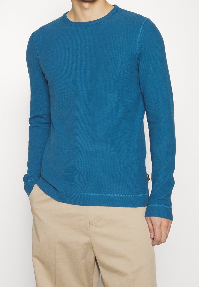 Blå långärmad tröja med rund hals i ribbad struktur, försedd med rak fåll och subtila sömnaddetaljer, kombinerad med beige byxor.