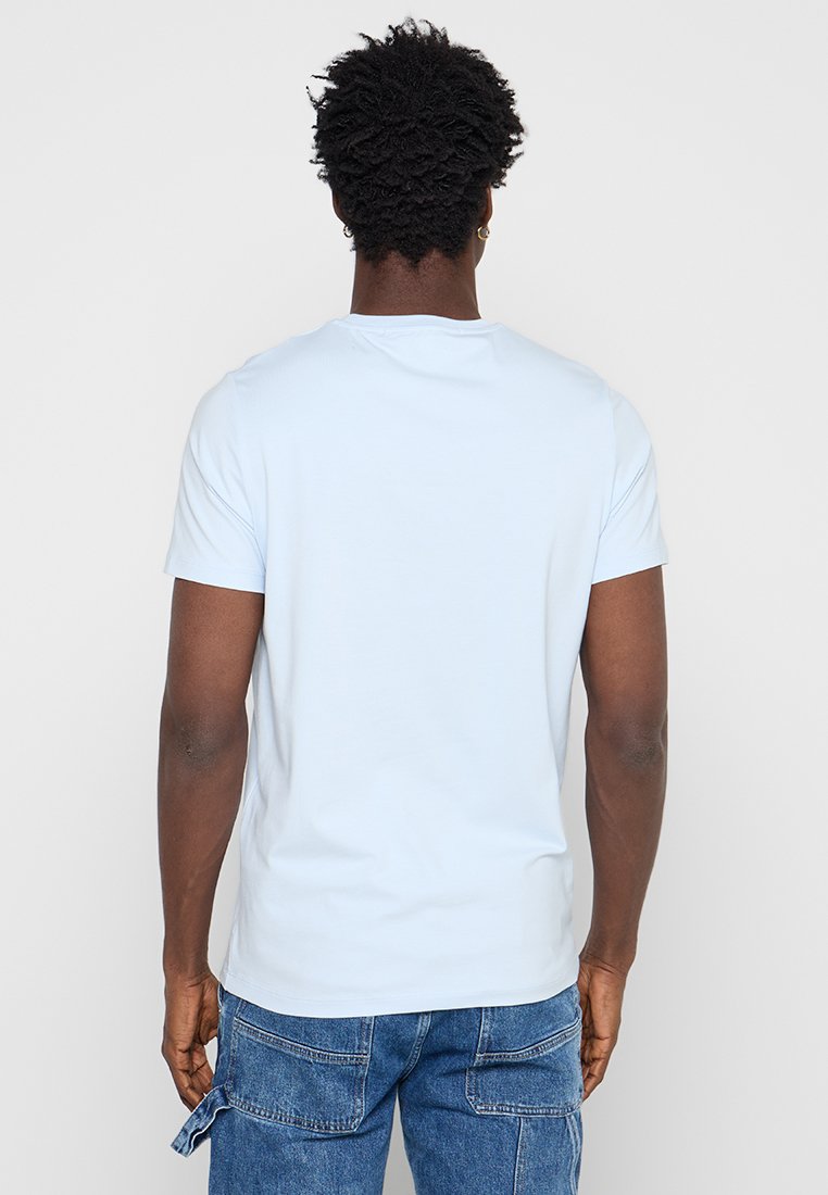 Hombre con pelo rizado y corto, vestido con una camiseta lisa de manga corta de color azul claro y pantalones vaqueros azules, de pie de espaldas frente a un fondo blanco.