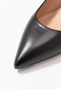 Sorte lædersko med spids tå og glat tekstur, der har en beige indvendig og en subtil syning langs kanten.