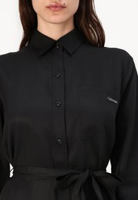 Calvin Klein svart skjortklänning med krage, långa ärmar, framknappar, bröstficka och midjeband. Slät tyg med en matt yta.