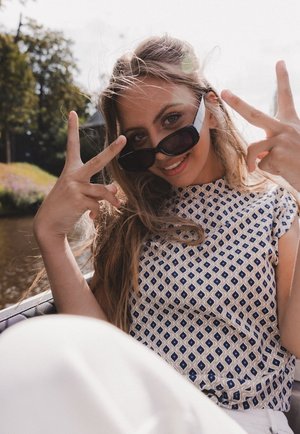 A patterned top with a blue geometric design, light fabric, and short sleeves; the person wears dark sunglasses and shows a peace sign.