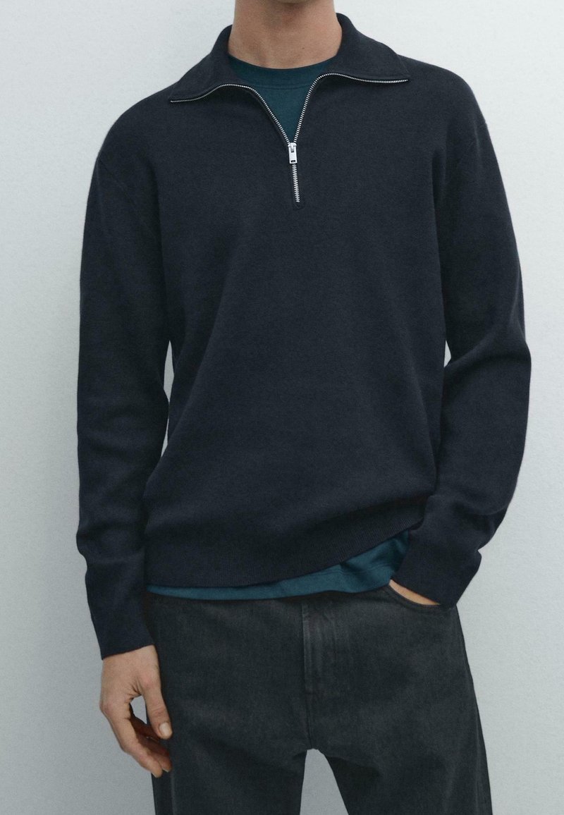Homme portant un pull à fermeture éclair bleu marine foncé par-dessus une chemise turquoise avec un pantalon noir, une main dans la poche, sur un fond clair uni.