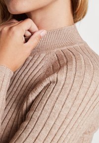 Beige gerippter Pullover mit strukturiertem Strick, der einen hohen Kragen und lange Ärmel hat. Eine Nahaufnahme zeigt die Weichheit des Materials.