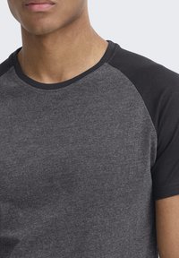 Camiseta gris con mangas raglán negras. Fabricada con una suave tela texturada. Presenta un clásico cuello redondo y un corte ajustado.
