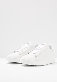 Scarpe sportive bianche in pelle, con una texture liscia, punta tonda, chiusura frontale con lacci e un marchio discreto sul lato.