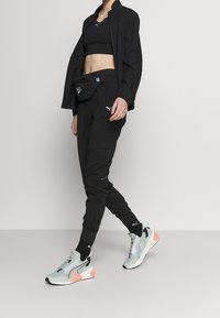 Schwarzes Sportoutfit mit einer cropped Jacke und hoch taillierter Hose, kombiniert mit hellgrauen Sneakers und schwarzen Socken. Akzente mit Puma-Logo.