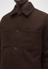 Homme portant une veste marron foncé boutonnée avec deux poches poitrine et un col pointu.