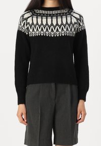 Svart stickad tröja med vitt geometriskt mönster vid oket, rund hals och ribbade muddar. Kombination med grå shorts för kontrast.