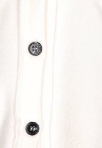 Fleece-tröja med en mjuk, krämfärgad textur som har svarta knäppningar och vertikala sömnadsdetaljer.