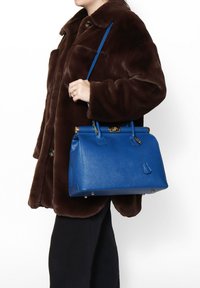 Gestructureerde handtas van gestructuurd blauwe leer, met gouden hardware, dubbele handvatten en een rechthoekige vorm, vastgehouden naast een bruine bontjas.