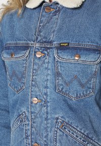 Blauwe denimjack met een schapenvachtkraag, koperen knopen, dubbele zakken en kenmerkende stikdetails in een zigzagpatroon.