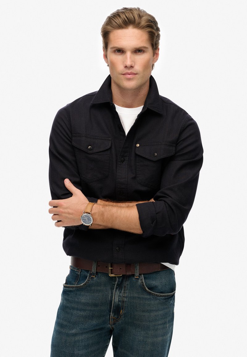 Zwart overhemd met knoopsluiting en twee borstzakken, gedragen over een wit T-shirt. Gecombineerd met blauwe jeans en een bruine leren horloge. Casual styling.
