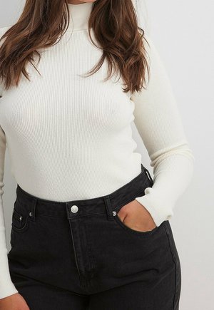 Femme portant un pull à col roulé côtelé blanc à manches longues et un jean noir taille haute, main dans la poche.