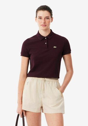 Vrouw draagt een donkerpaarse polo en beige short met koordsluiting, staat met één hand in de zak en met de andere hand een tashandvat vasthoudend.