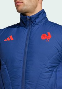 Blå zip-up jacka med hög krage, prydd med röda detaljer inklusive en tupp-logotyp och diagonala ränder på vänster axel. Strukturerad tyg.
