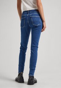Jeans de mezclilla azul, ajustados y de cintura alta, con bolsillos traseros y detalles de costura, combinados con botas negras hasta el tobillo, de pie sobre un fondo neutro.