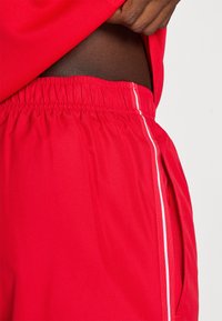 Rdečkasti športni kratki hlače iz lahkotnega, zračnega materiala, s teksturirano površino, elastičnim pasom in belimi stranskimi črtami.