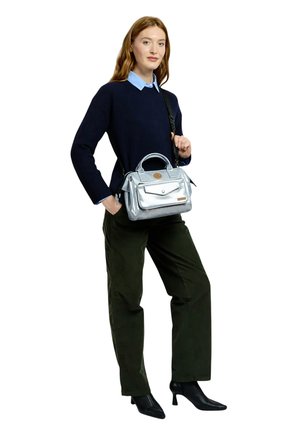 Femme debout, main dans la poche, portant un pantalon noir, un pull marine par-dessus une chemise bleue, des talons noirs, et tenant un sac à main argenté avec une bandoulière.