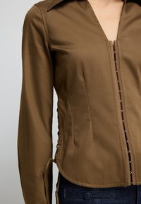 Brown long-sleeve fitted blouse with hook-and-eye front closure and side lace-up detail worn with dark blue jeans.
