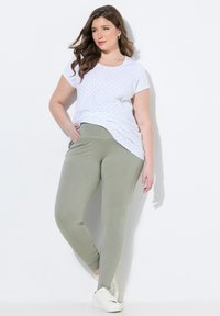 Bílý krátký rukáv tričko s malými černými tečkami a olivově zelené legíny s kapsami, nošené s bílými teniskami. Hladká struktura látky.