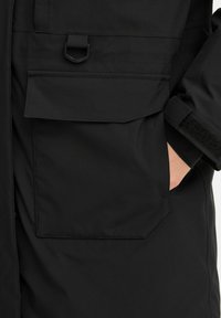 Čierny nepremokavý kabát s veľkou prednou kapsou, hrubou tkaninou a uzávermi na suchý zips na rukávoch. Hladká textúra s minimálnymi švovými detailmi.