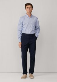 Joven de pie contra un fondo liso vistiendo una camisa de botones a rayas azul claro, pantalones oscuros de color navy y zapatos beige.