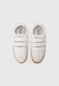 White sneakers with three textured Velcro straps, smooth leather and suede materials, gum rubber sole, centered logo on the tongue.
