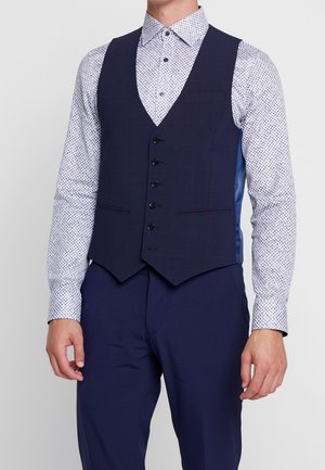 Uomo che indossa un gilet blu navy con bottoni sopra una camicia bianca a maniche lunghe con motivo e pantaloni blu navy abbinati.