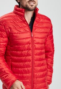 Chaqueta acolchada roja con diseño de acolchado horizontal, cuello alto y cremallera completa. El material parece brillante y ligero. Sin patrones visibles.