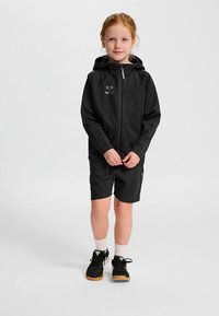 Svart zip-up hoodie med huva, kombinerad med svarta shorts. Barnet bär vita strumpor och svarta sneakers med beige sulor. Hoodien har en logotyp.