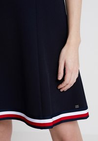 Vestido azul marino con un diseño acampanado, que presenta un dobladillo a rayas en rojo, blanco y azul. Incluye una pequeña etiqueta con el logo cerca de la parte inferior.