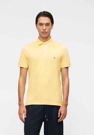 Giovane uomo in piedi, indossa una polo gialla a maniche corte e pantaloni neri su uno sfondo chiaro e uniforme.