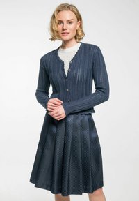 Marineblauer Rippstrick-Cardigan mit Knopfschluss, kombiniert mit einem plissierten, gemusterten marineblauen Rock. Strukturiertes Strickmaterial und glattes Gewebe betonen das Design.