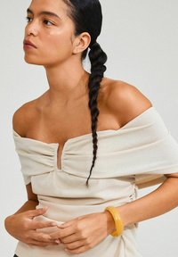 Off-shoulder, ljusbeige topp med rynkad frontdesign, kombinerad med ett gult armband. Slätt texturerat tyg.