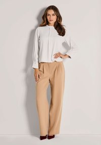 Frau mit langen, welligen Haaren trägt eine weiße Bluse mit langen Ärmeln, beigen, weiten Hosen und dunkell roten, spitz zulaufenden Schuhen und posiert vor einer hellen Wand.