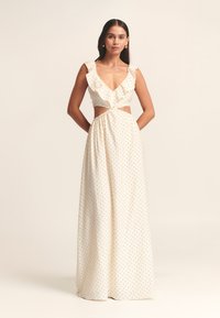 Vestido largo de color crema con lunares negros. Presenta un escote en V profundo, mangas con volantes, un corte frontal y una falda fruncida para textura.