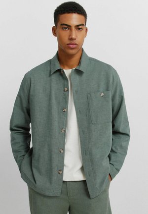 Młody mężczyzna z krótkimi kręconymi włosami, ubrany w zieloną kurtkę na guziki na jasnym odcieniu szałwii, z białą koszulą pod spodem, trzymający ręce w kieszeniach.