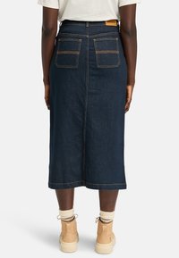 Mørkeblå denim midi-nederdel med lige snit, to baglommer, kontrasterende syninger og et lædermærke ved taljen.