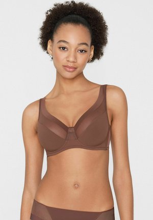 Žena se zvlněnými vlasy, oblečená v hnědé podprsence s kosticemi a odpovídajícím spodním prádle, stojící před jednoduchým světlým pozadím.