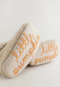 Beige neulejalkineet, joissa on tekstuurinen kuviointi, ja jotka sisältävät oranssia "pikkupumpuli" brodeerausta pohjissa lisäyksinä.