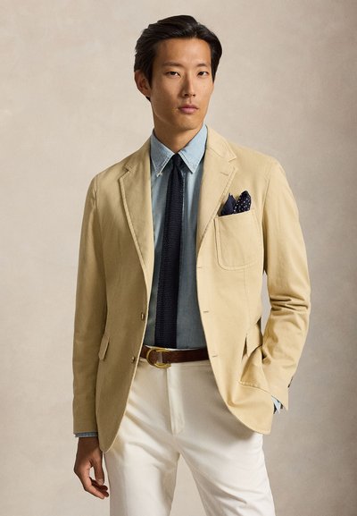 Homme portant un blazer beige, une chemise bleu clair, une cravate noire, un pantalon blanc et une ceinture marron, la main dans la poche sur fond neutre.
