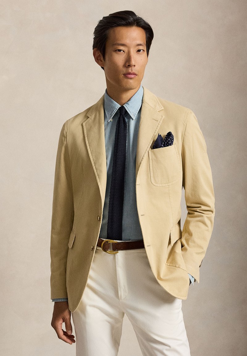 Homme portant un blazer beige, une chemise bleu clair, une cravate noire, un pantalon blanc et une ceinture marron, la main dans la poche sur fond neutre.