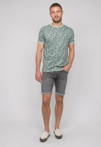 T-shirt vert clair à manches courtes avec un motif de feuilles et une bordure blanche, associé à un short gris retroussé et des chaussures beiges à enfiler.