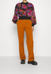 Pull à motifs multicolores avec manches bouffantes et bordures côtelées noires, associé à un pantalon orange ample et des chaussures noires épaisses.