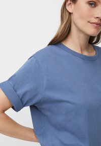 Žena s dlouhými světlými hnědými vlasy, oblečená v modrém tričku s krátkým rukávem, ruka opřená o bok, na jednoduchém bílém pozadí.