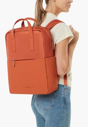 Woman wearing a square, burnt orange backpack with top handles, front zipper pocket, and padded shoulder straps, paired with a short-sleeve sweater and jeans.